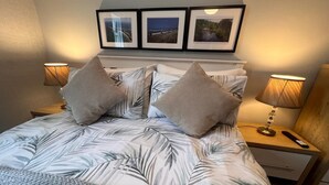 Una cama bien hecha con una colcha estampada con motivos tropicales, dos almohadas a juego, una lámpara de noche y tres cuadros enmarcados en la pared de arriba.
