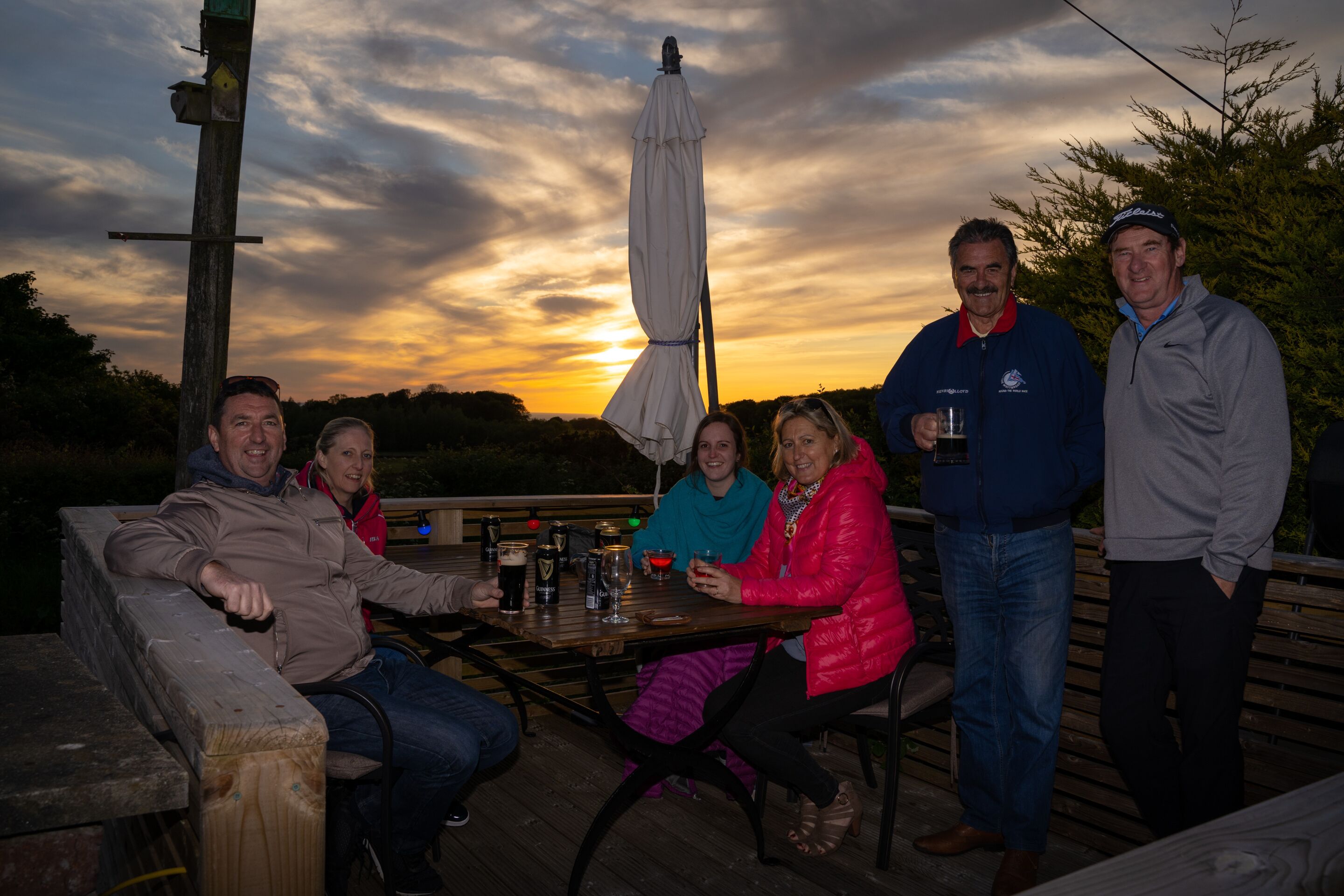 A group of people seated at a wooden table outdoors with a sunset in the background.