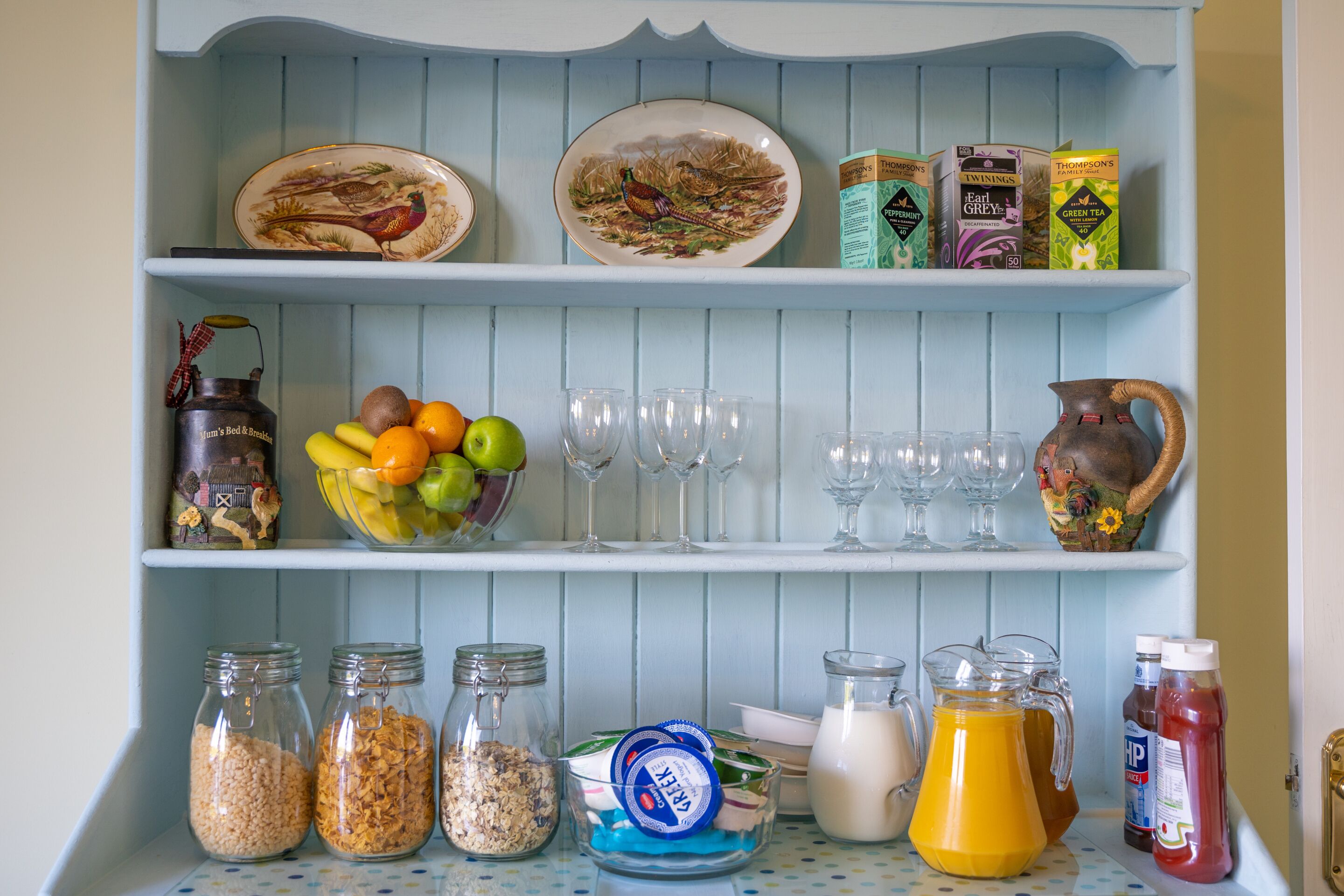 A kitchen shelf with decorative plates, a fruit bowl, glassware, and food storage containers.