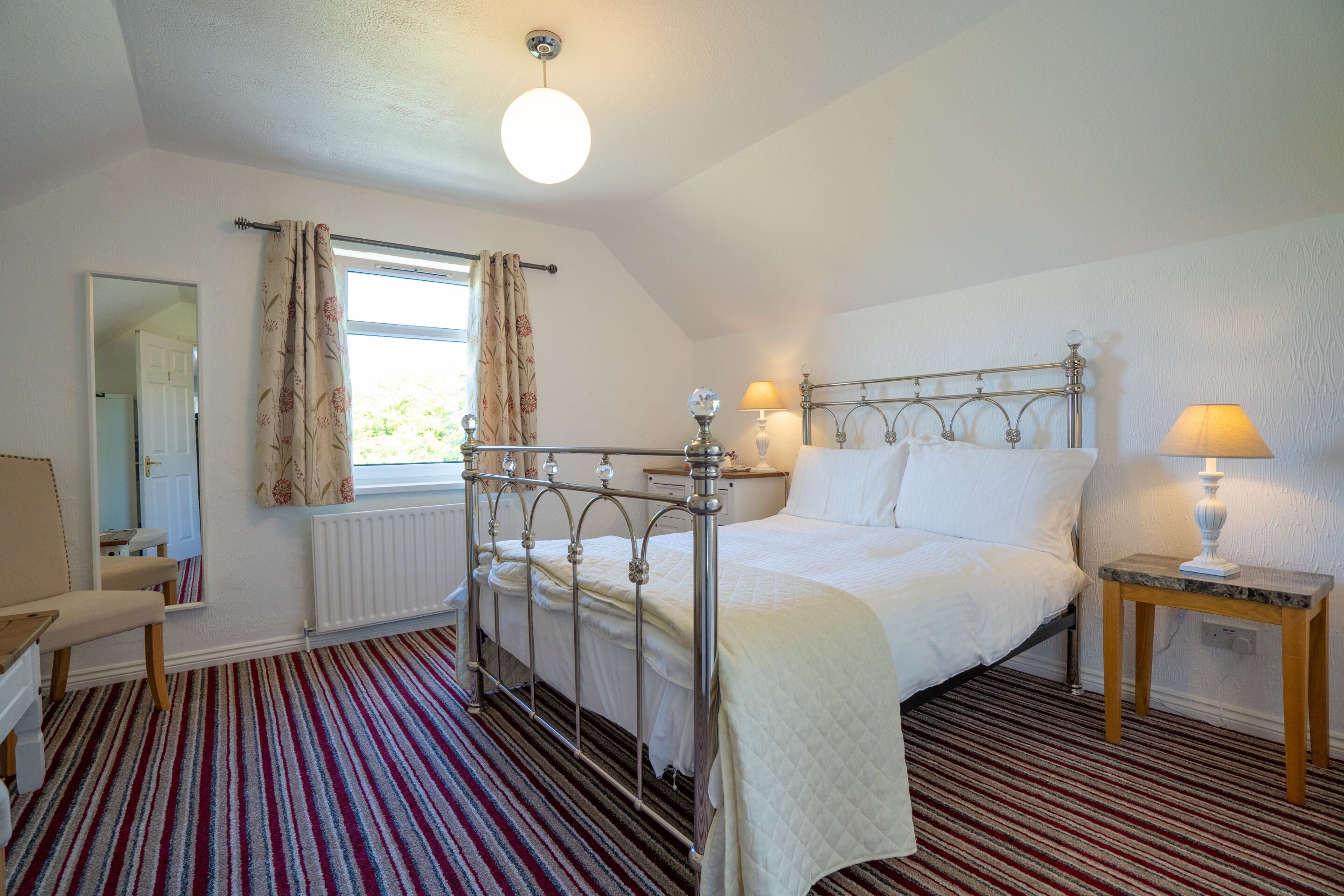 A bedroom with a metal bed frame, two bedside lamps, a window with curtains, a radiator, and a striped carpet.