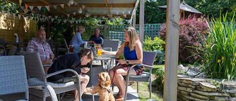 A covered outdoor seating area with wicker chairs and tables, surrounded by greenery and a stone wall.