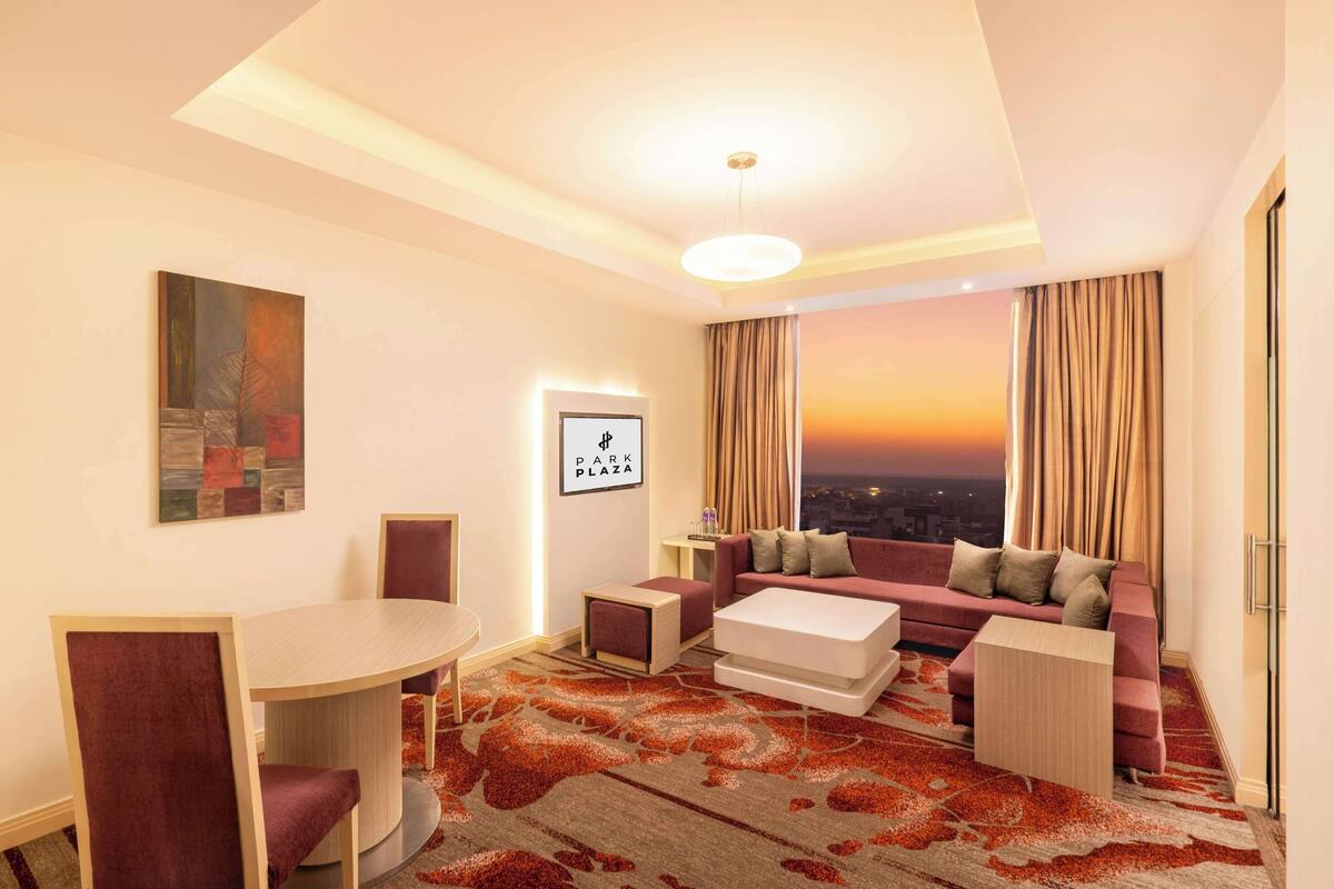 Habitación de hotel moderna con zona de comedor, conjunto de sofá y vista a la ciudad al atardecer.