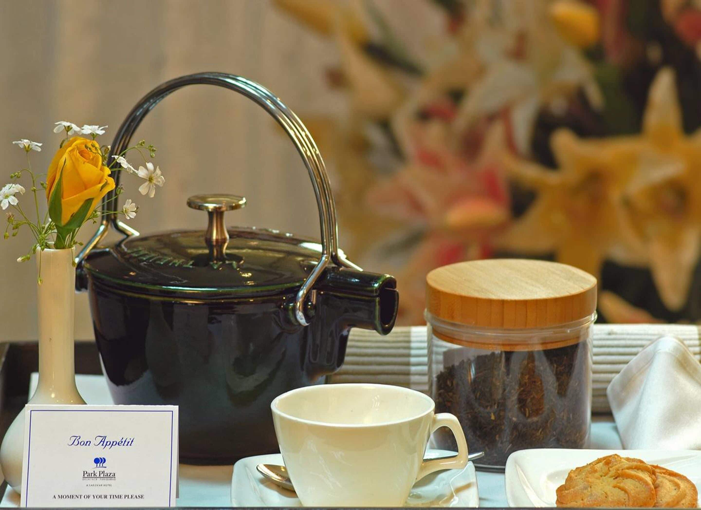 A tea set with a black teapot, a white cup, a glass jar with a wooden lid, and a plate with cookies. A vase with a yellow rose and small white flowers is also visible.