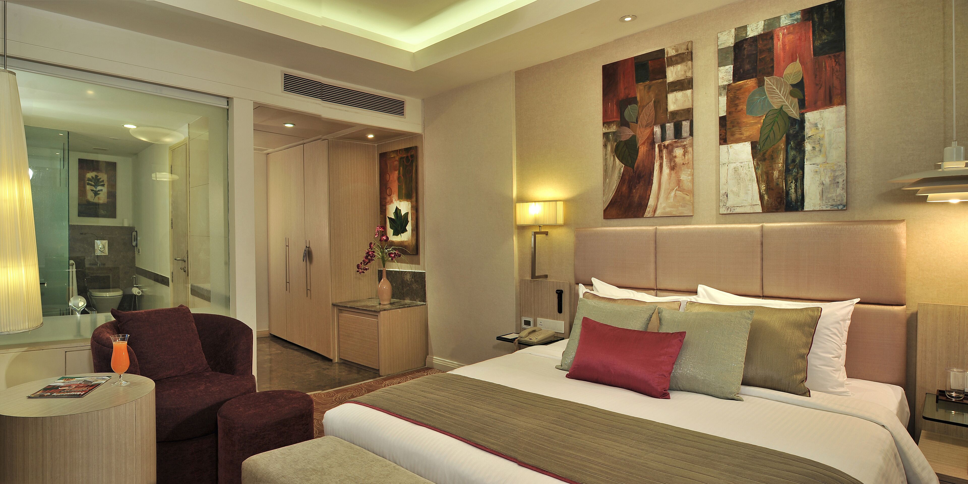 Une chambre d’hôtel avec un grand lit, un coin salon avec un fauteuil, un petit bureau et une salle de bain visible à travers une porte ouverte.