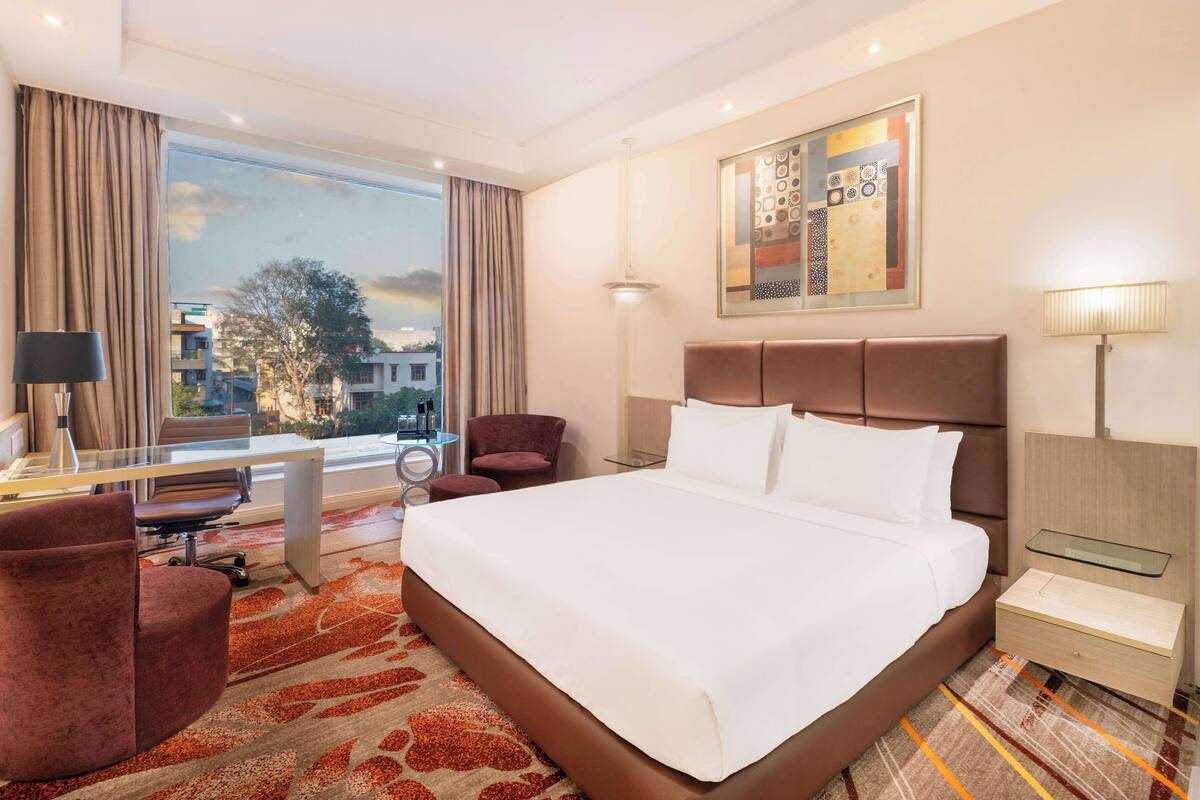 Habitación de hotel con una cama grande, un escritorio con una silla, un sillón pequeño, una mesita de noche y una lámpara. Hay una ventana con cortinas, un cuadro en la pared y vistas de edificios.