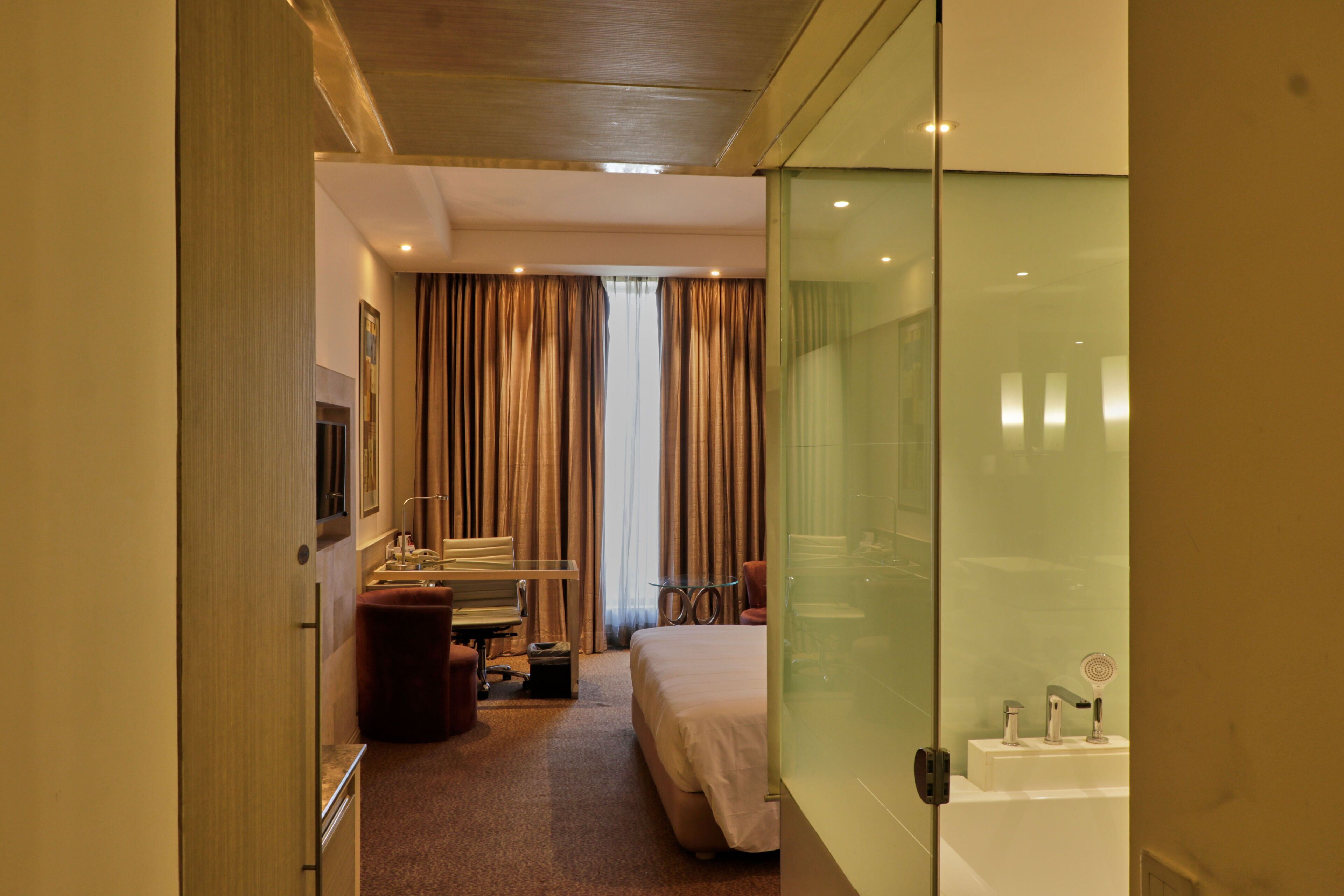 A hotel room with a bed, a desk, a chair, a small table, a mirror, and a bathroom with a sink and a towel rack.