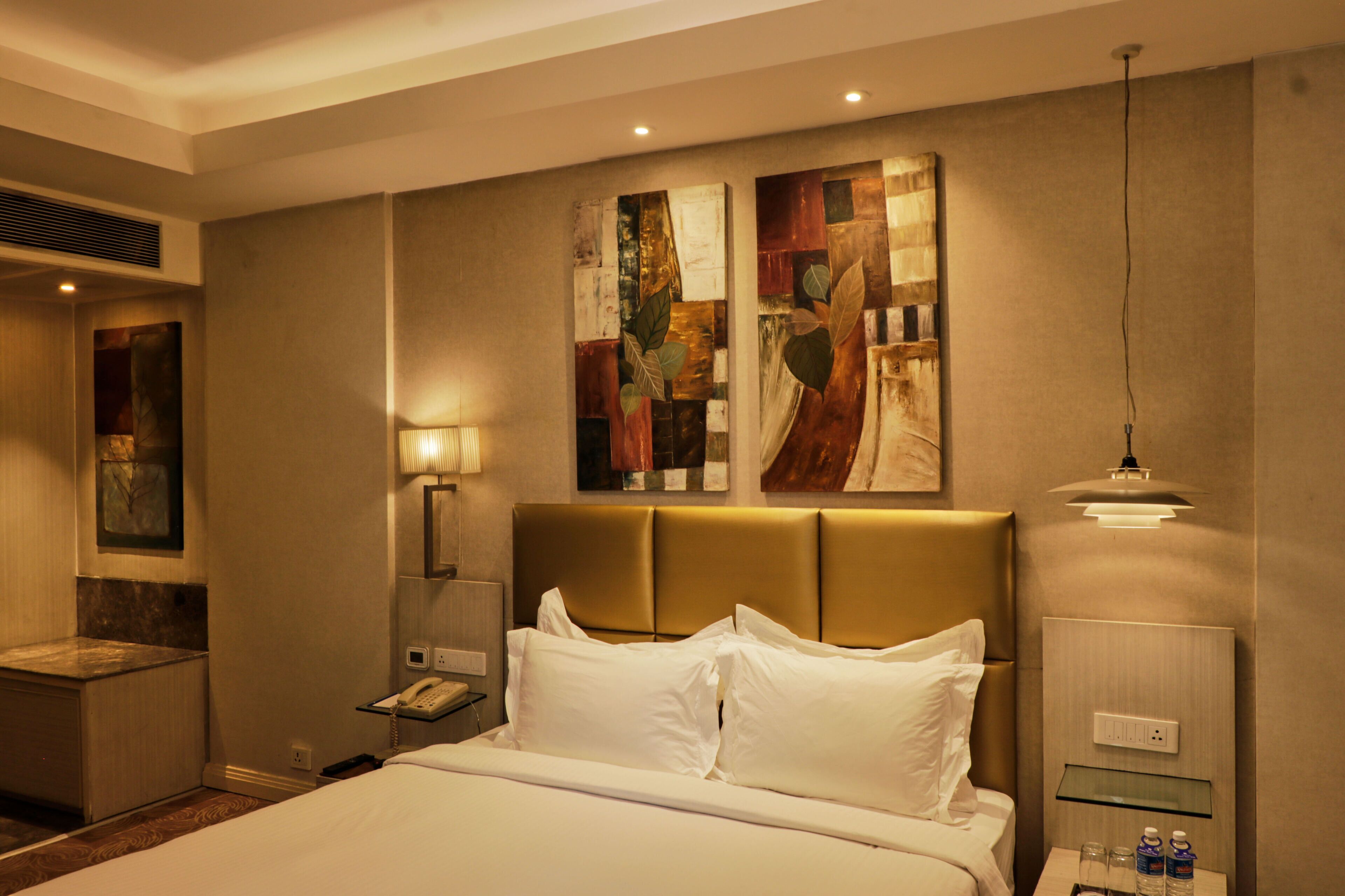 A hotel room with a large bed, a bedside table with a lamp, a phone, and a wall-mounted shelf with bottles.