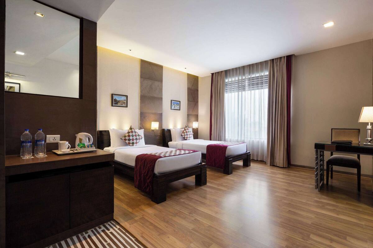 Une chambre d’hôtel avec deux lits, un bureau, une chaise, un miroir et une fenêtre avec des rideaux.