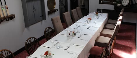 Una larga mesa de comedor con manteles blancos, cubiertos y cristalería, flanqueada por sillas de madera y sillones tapizados de rojo.