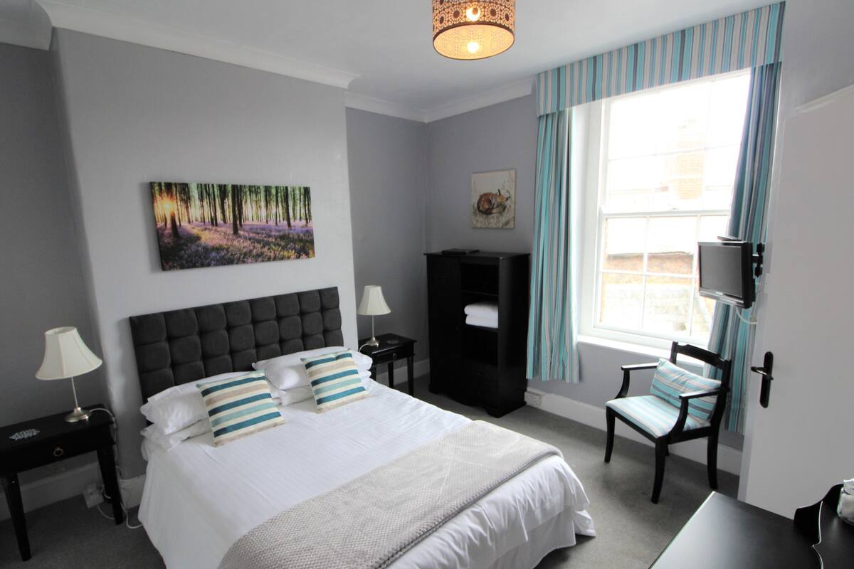 A neatly arranged hotel room with a large bed, a bedside table with a lamp, a TV mounted on the wall, and a window with curtains.