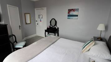 Ein Schlafzimmer mit einem großen Bett, einem Schminktisch, einem Stuhl, einem Schreibtisch mit Spiegel und einem Bild an der Wand.