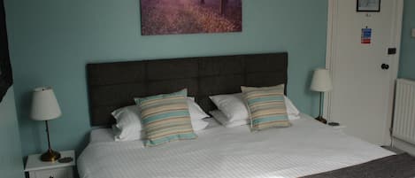 A neatly made bed with two pillows and a blanket, a bedside lamp, a framed picture on the wall, and a door with a fire action sign.