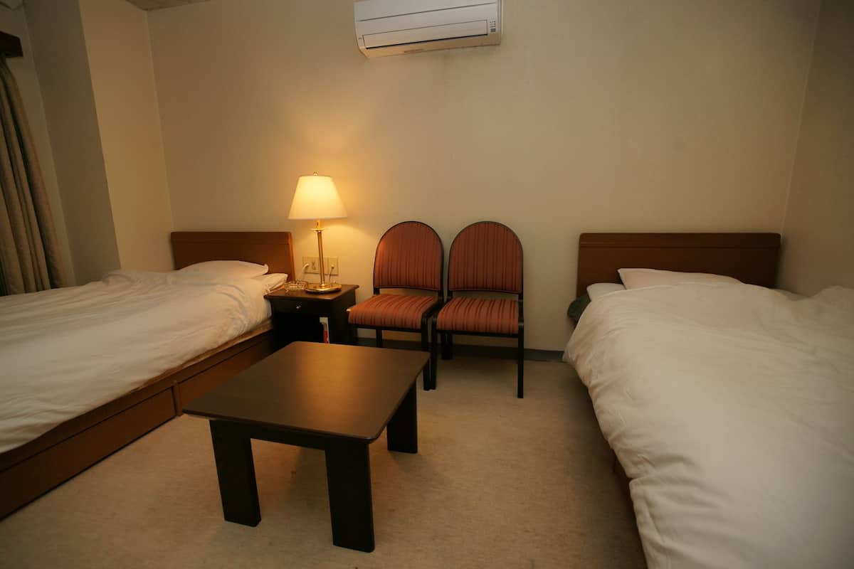 A hotel room with two single beds, a small table, two chairs, a lamp, and an air conditioning unit.