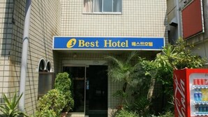 The exterior of Best Hotel with a tiled facade, a Coca-Cola vending machine, and some greenery in front.