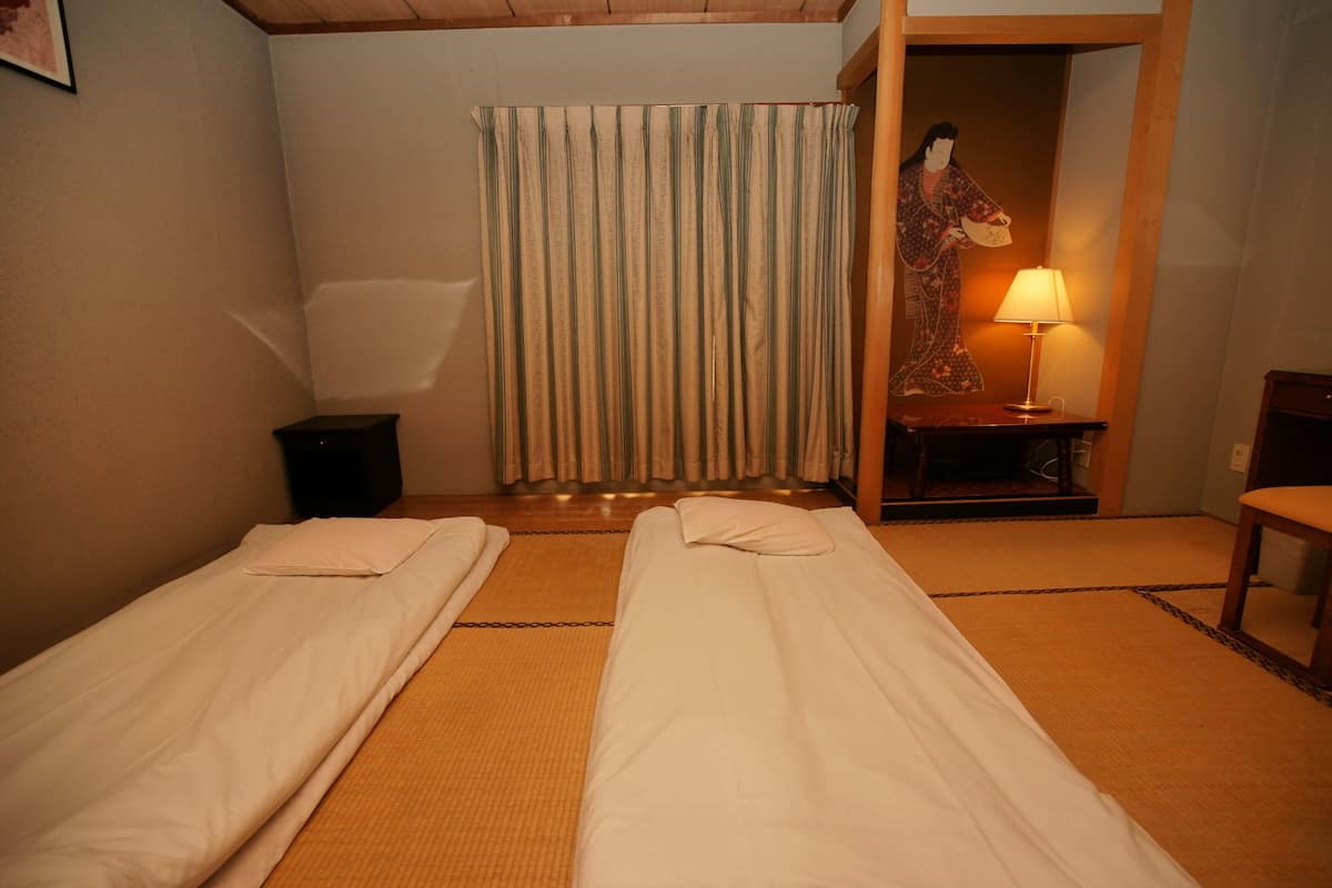 A room with two single beds, a wooden floor, a curtain, a small table with a lamp, and a wall-mounted picture.