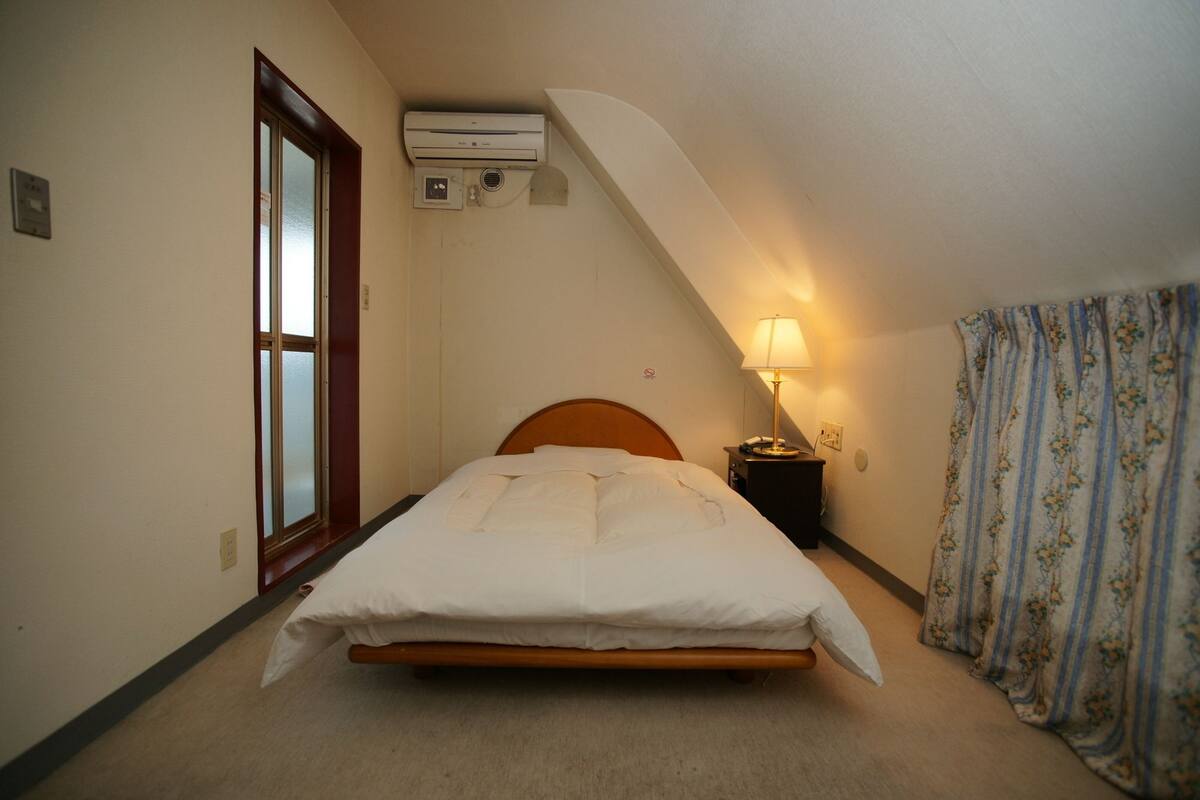 A single bed with a wooden headboard in a room with a sloped ceiling. There is a window with frosted glass, an air conditioning unit mounted on the wall, and a bedside lamp.