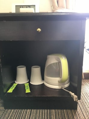Um armário preto com frente transparente, contendo uma chaleira elétrica verde e branca, duas xícaras de papel brancas e um objeto branco com etiqueta verde.