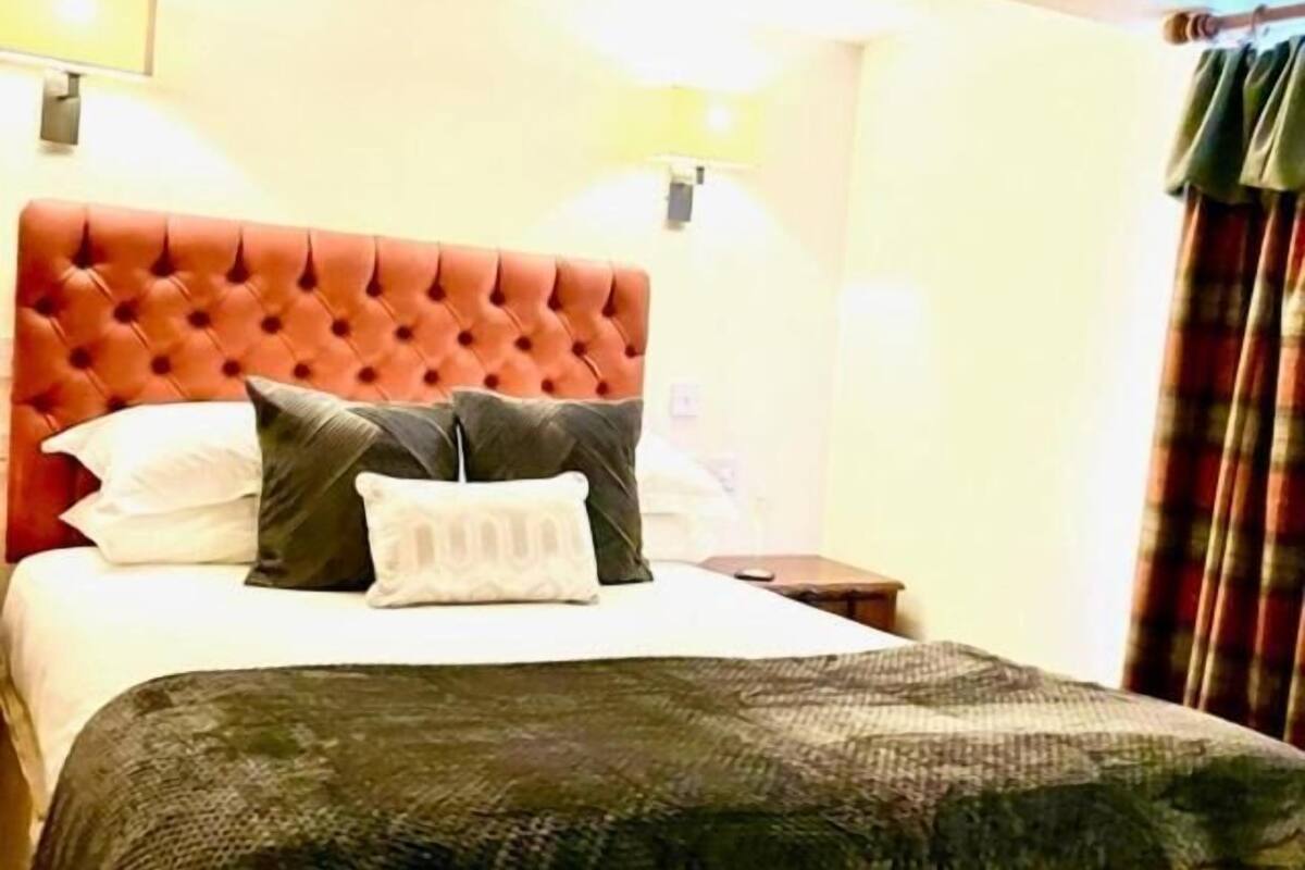 A hotel room with a tufted headboard, a bed with white and dark pillows, a patterned throw blanket, a bedside table, and a lamp.