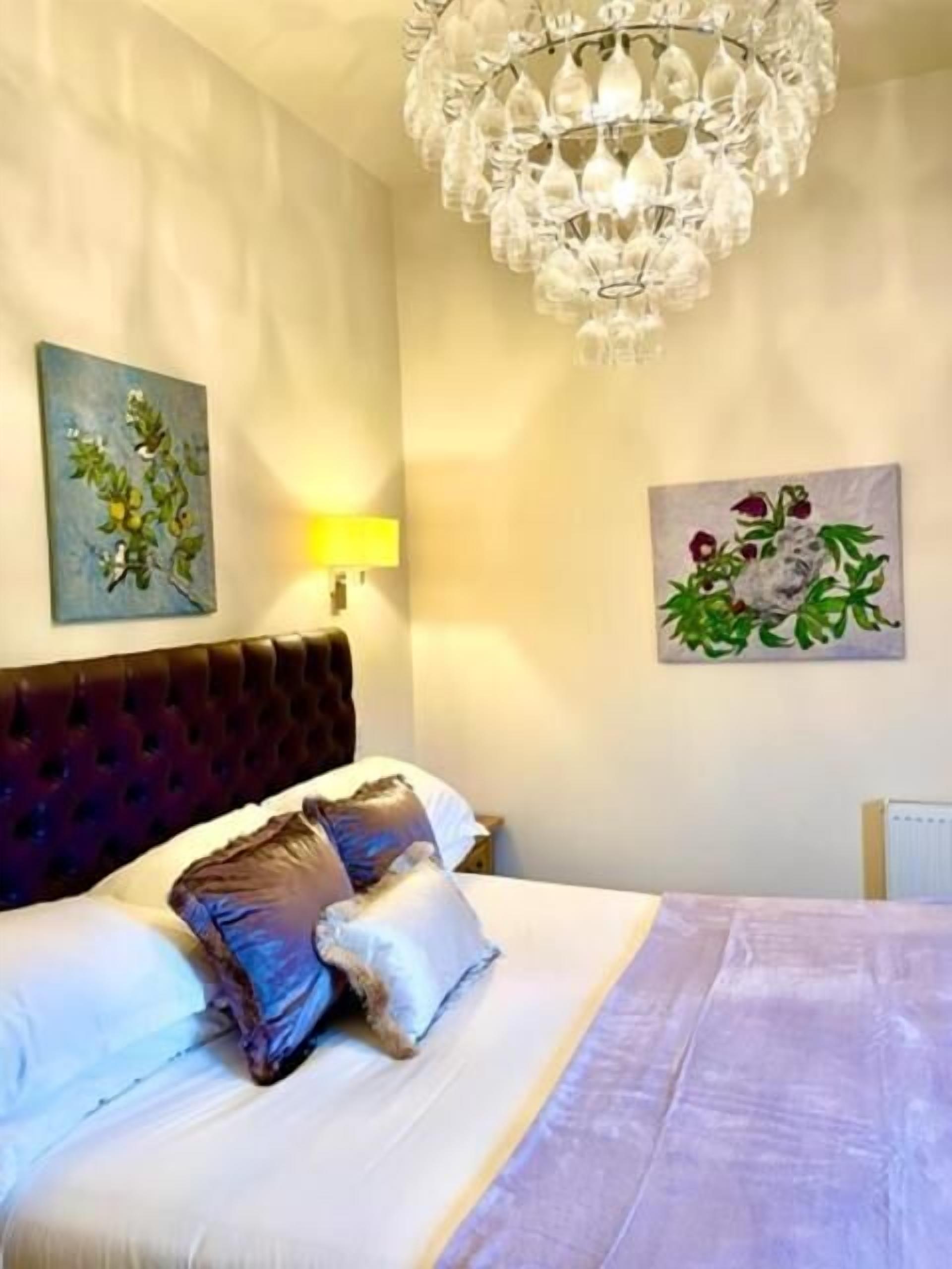 Une chambre à coucher avec une tête de lit capitonnée, un grand lit avec de nombreux oreillers, un lustre et deux tableaux accrochés au mur.