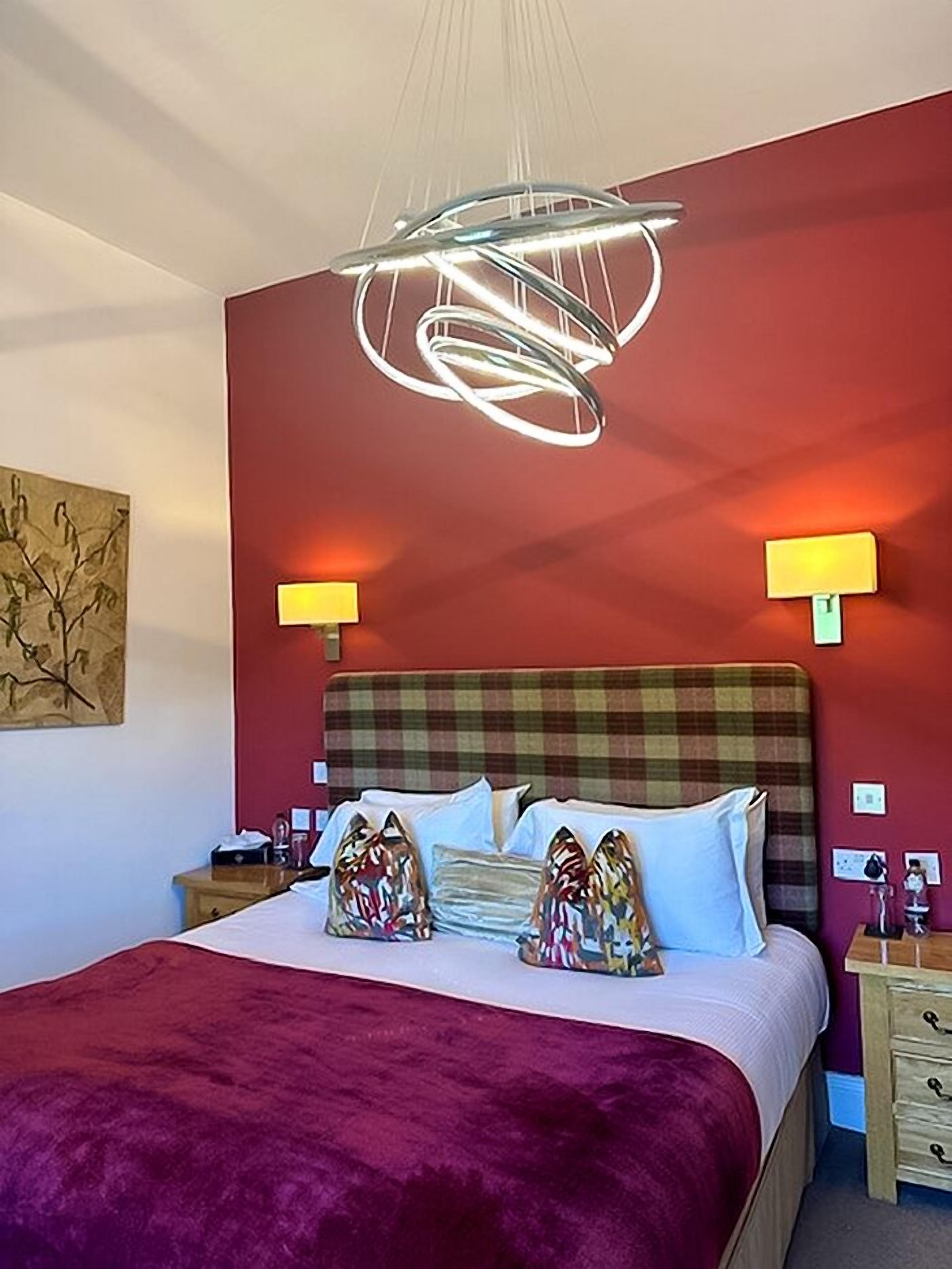 Een slaapkamer met een rode wand, een bed met witte beddengoed, een geruit hoofdeinde en twee bedlampjes.