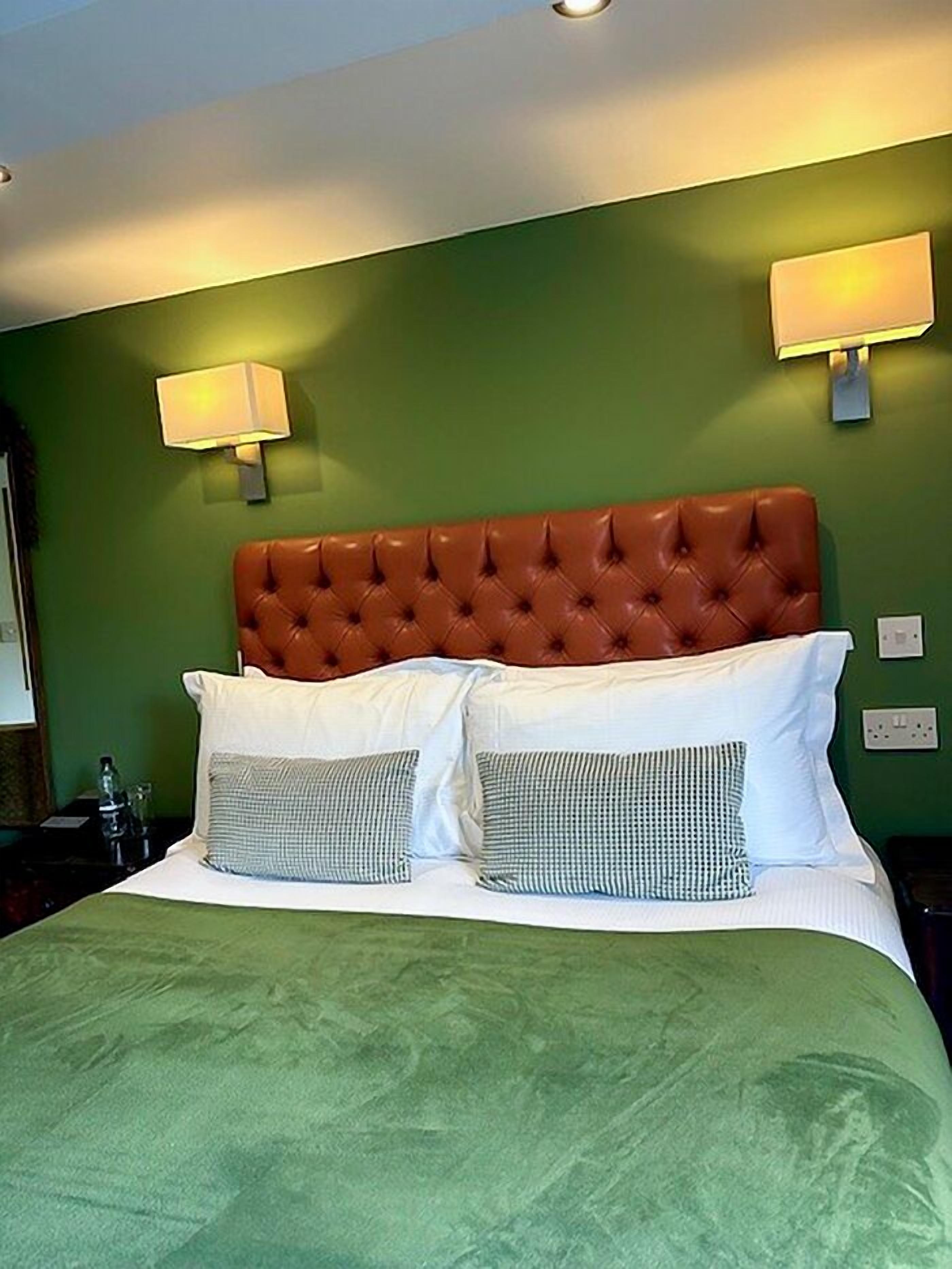 A hotel room with a green wall, a tufted brown headboard, white bedding, and two wall-mounted lamps.
