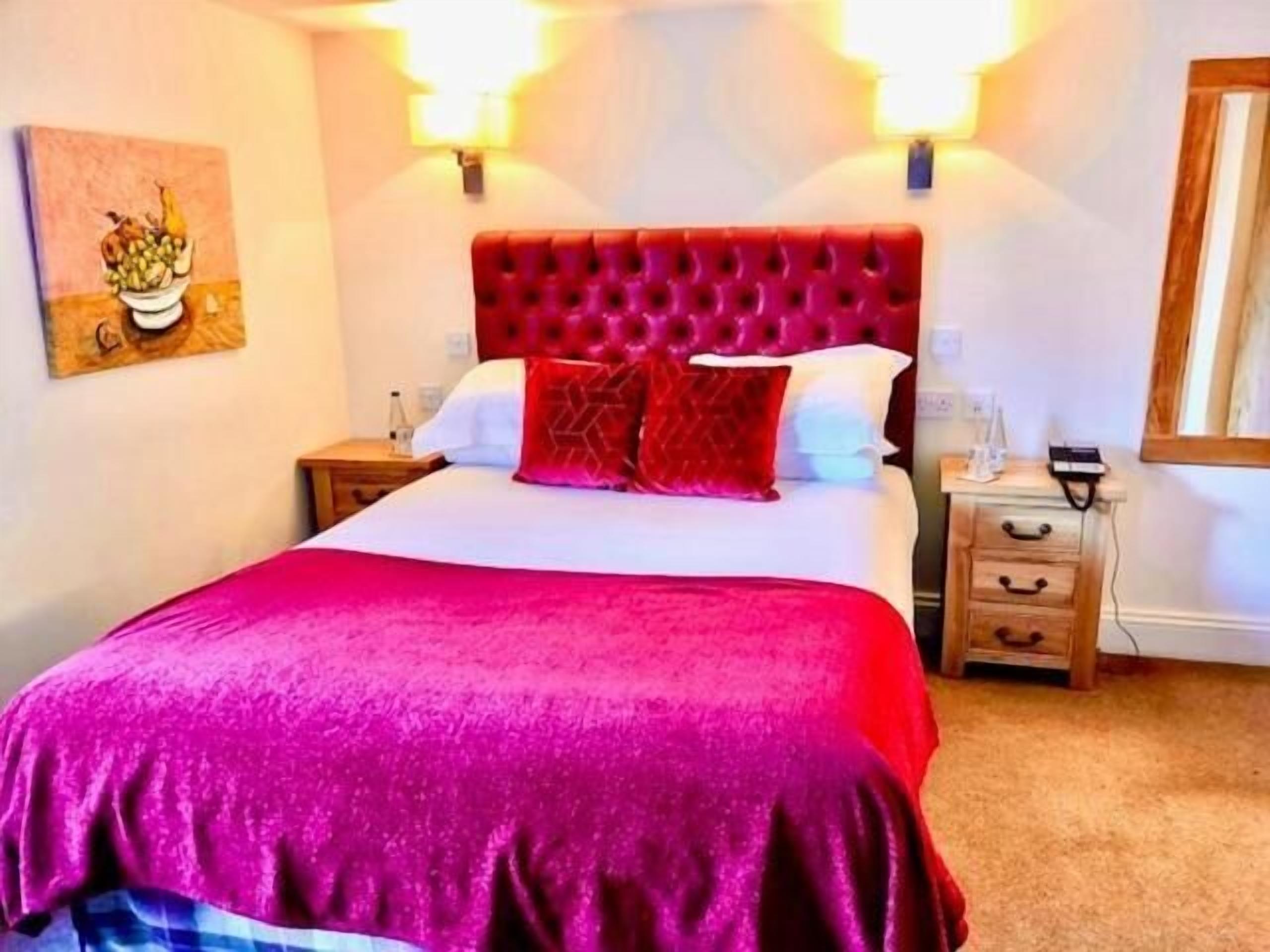 Quarto de hotel com uma cama grande, roupa de cama vermelha e branca, cabeceira estofada vermelha, mesas de cabeceira e um quadro na parede.