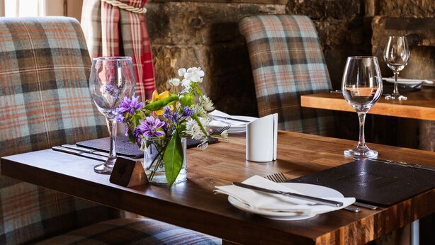 Une table de salle à manger en bois, agrémentée d’un vase de fleurs, de deux verres à vin et d’un chandelier, le tout placé devant un mur en pierre.