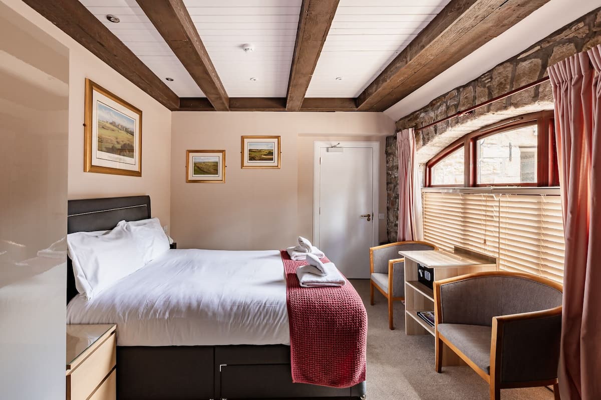 Un dormitorio con cama, mesita de noche, silla y un pequeño escritorio con asiento. Hay cuadros en la pared, una ventana con cortinas y una puerta.
