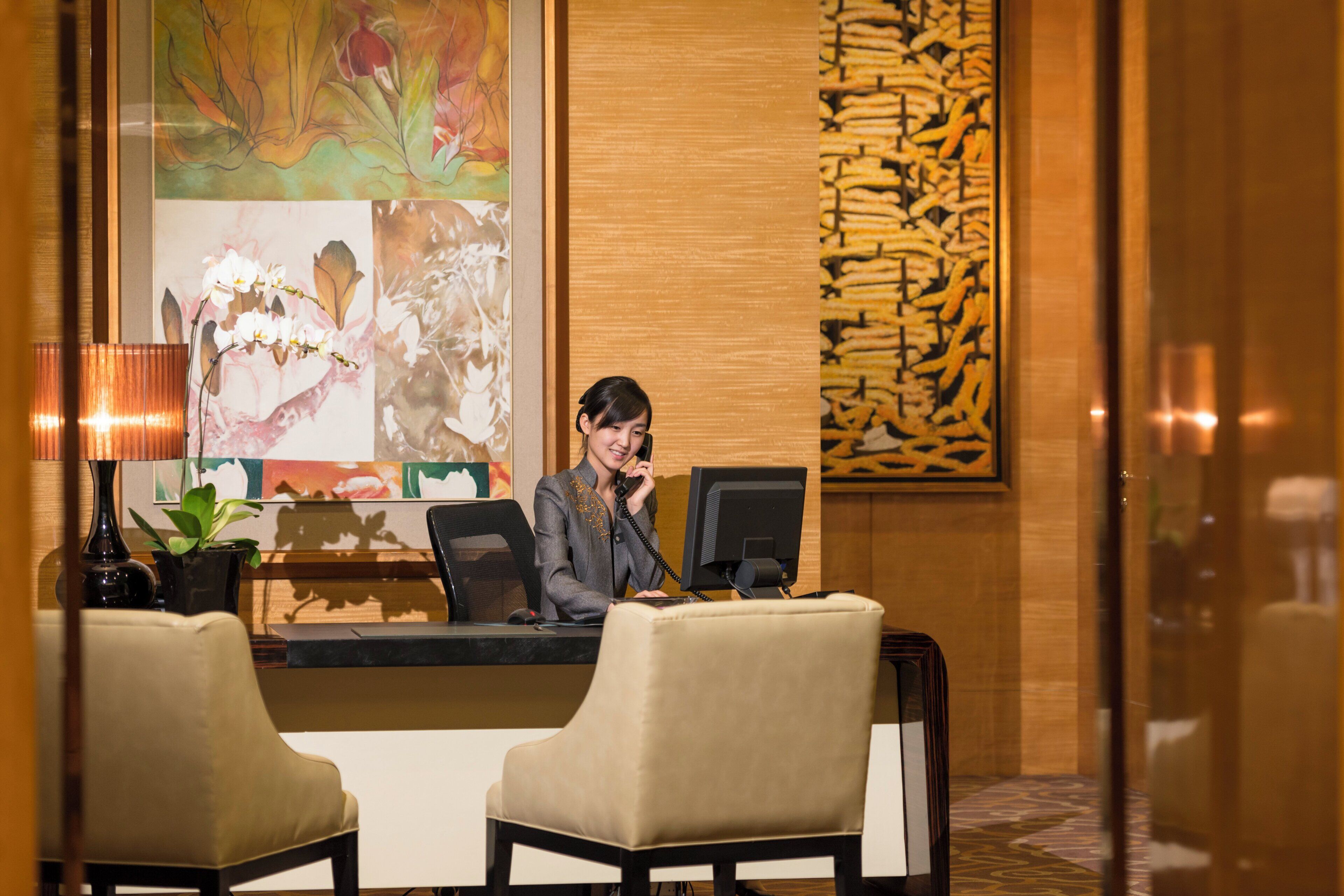 A reception desk with a computer, a telephone, and a chair. There is a plant on the desk and a lamp to the left.