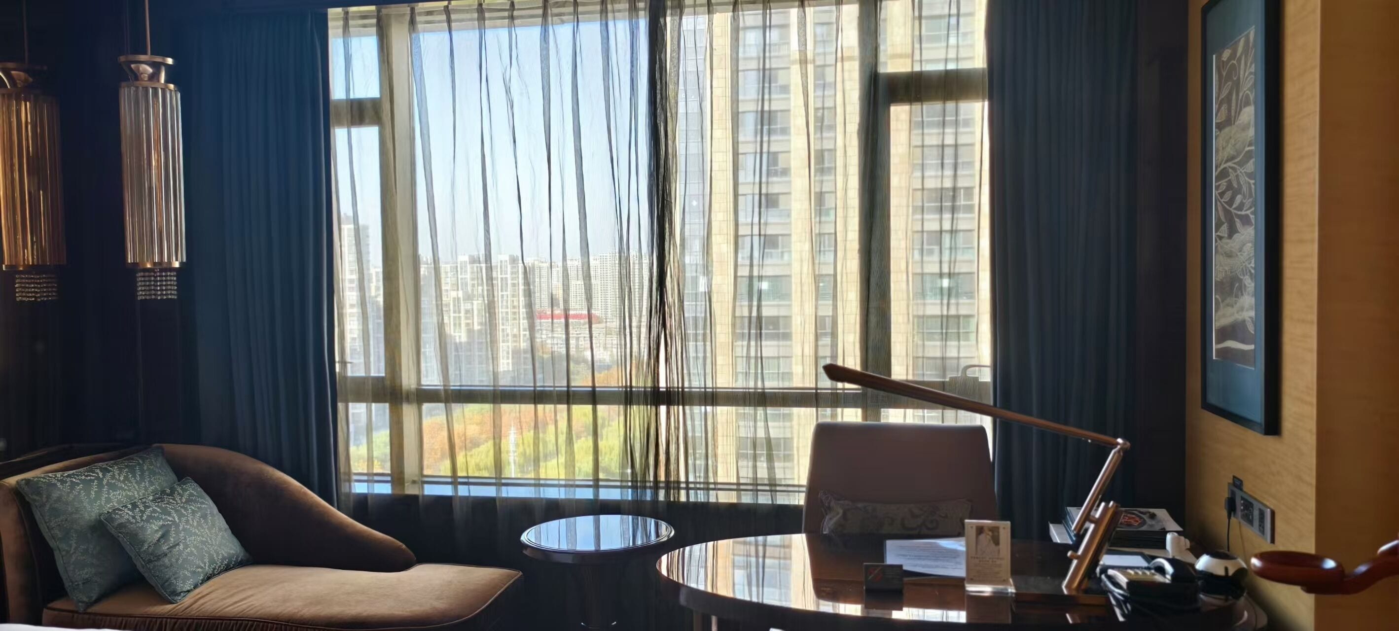 A modern hotel room with a large window, a brown leather sofa, a round table, and a desk with a chair.