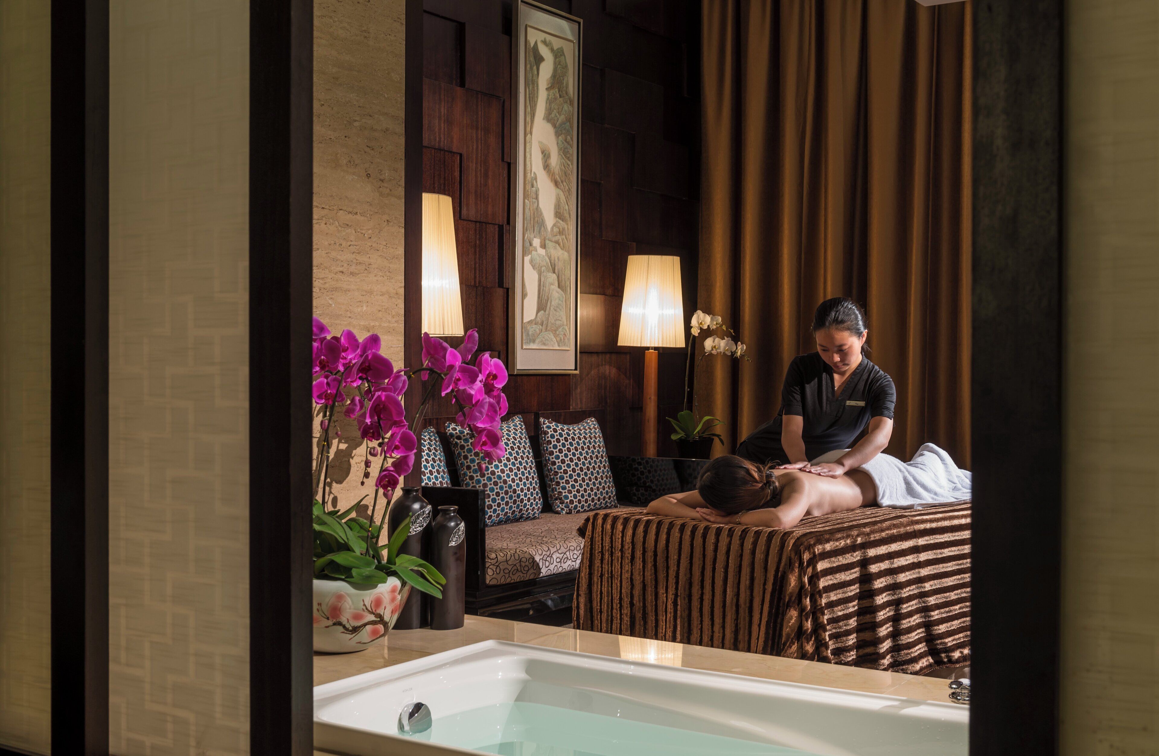 A spa room with a massage table, a massage therapist, and a bathtub.