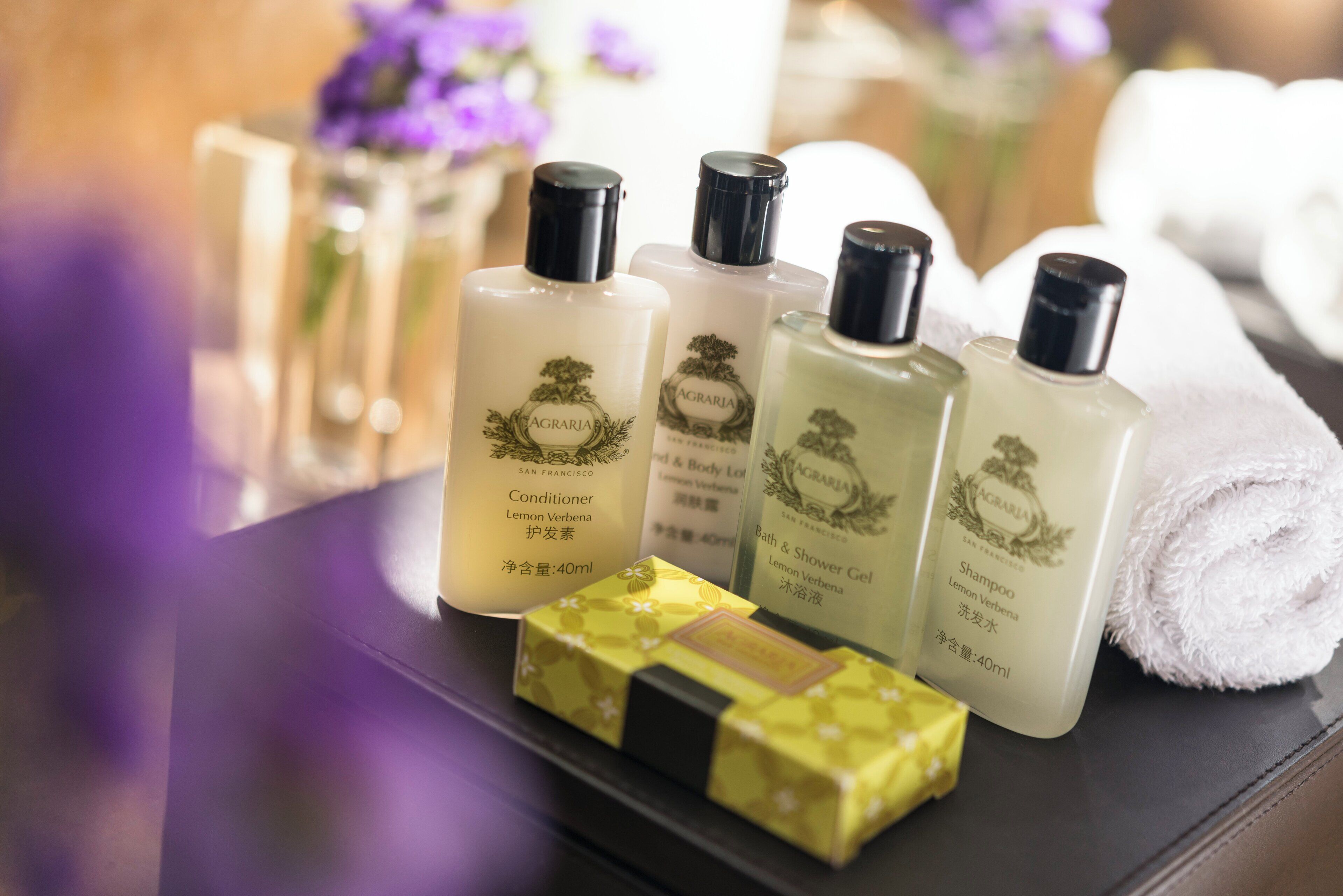 A set of toiletries including a conditioner, bath & shower gel, and shampoo, arranged on a table with a rolled towel and a decorative box.
