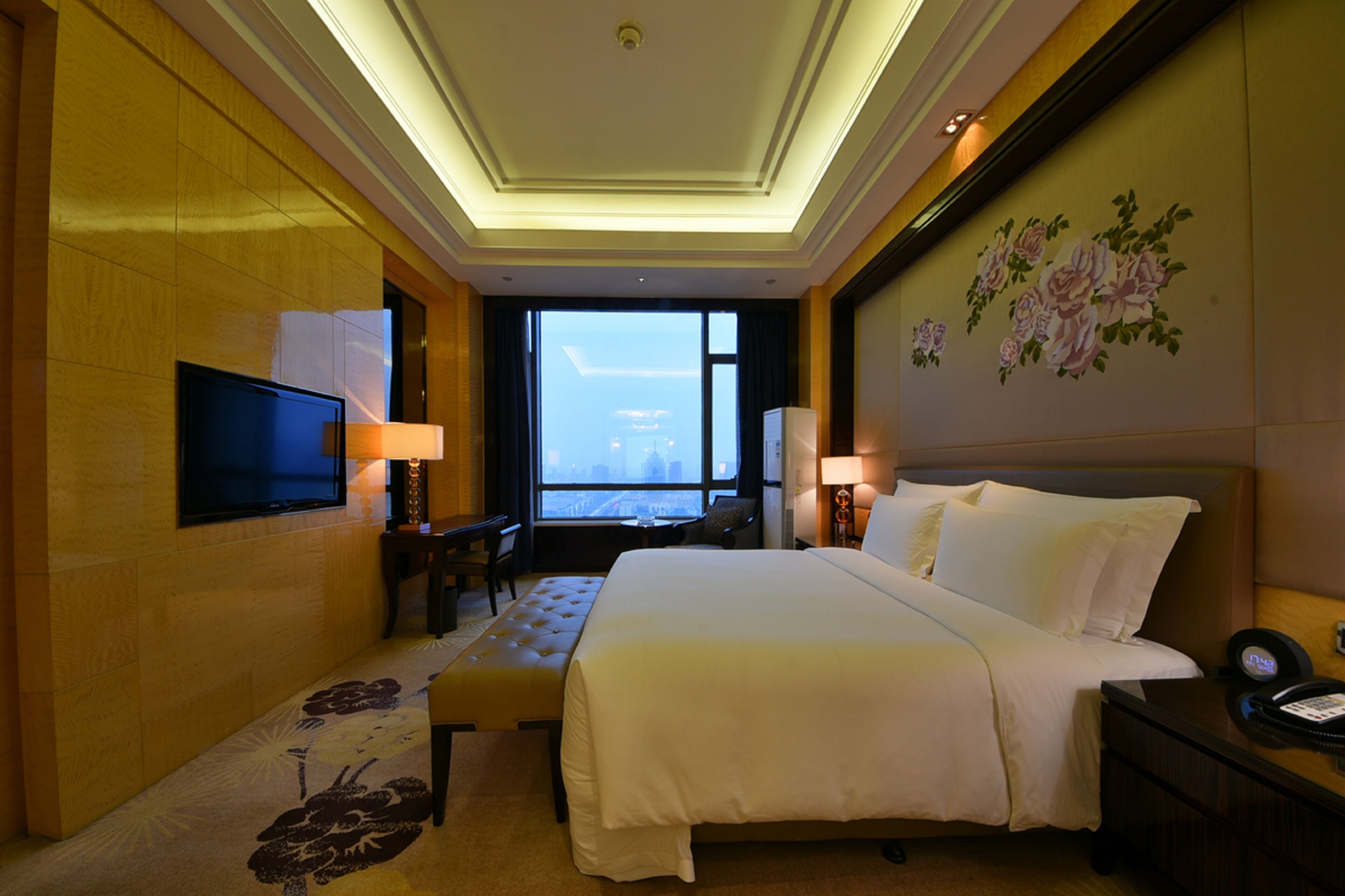Ein Hotelzimmer mit einem großen Bett, einem an der Wand befestigten Fernseher, einem Schreibtisch mit Lampe und Blick auf die Stadt durch das Fenster.