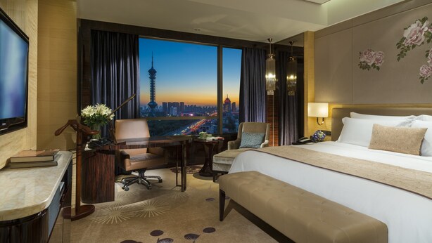 Habitación de hotel con una cama grande, un escritorio con silla, vista a la ciudad y una torre urbana.