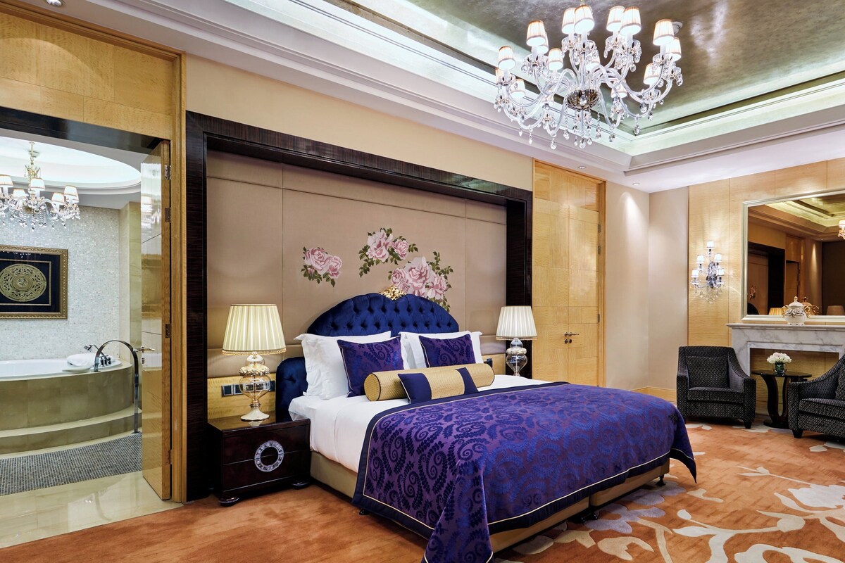 Een luxe hotelkamer met een groot bed, een zithoek met stoelen, een open haard en een decoratieve spiegel.