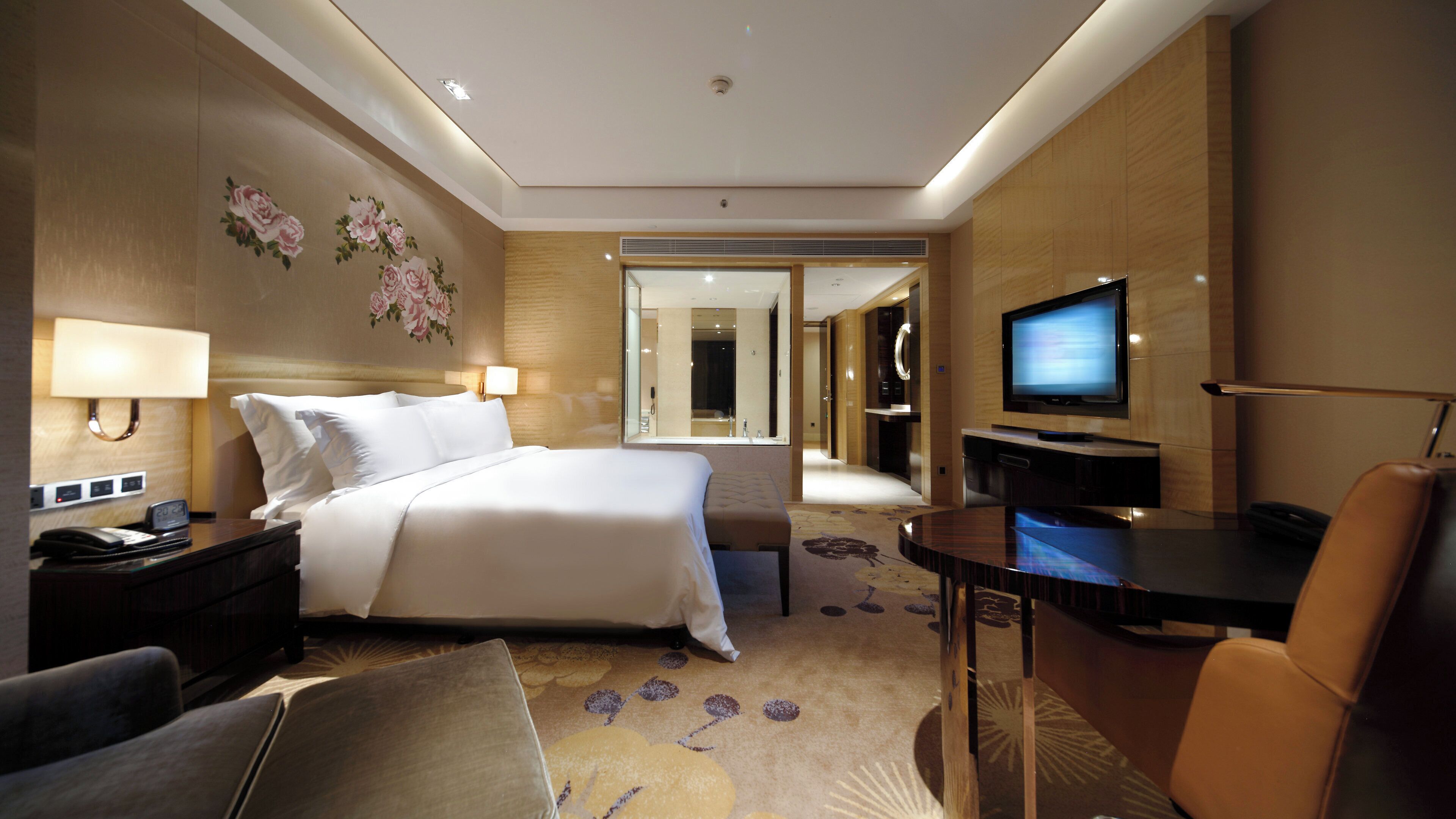 A hotel room with a bed, bedside tables, a desk, a TV, a chair, and a bathroom visible through a glass door.