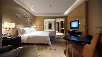 A hotel room with a bed, bedside tables, a desk, a TV, a chair, and a bathroom visible through a glass door.