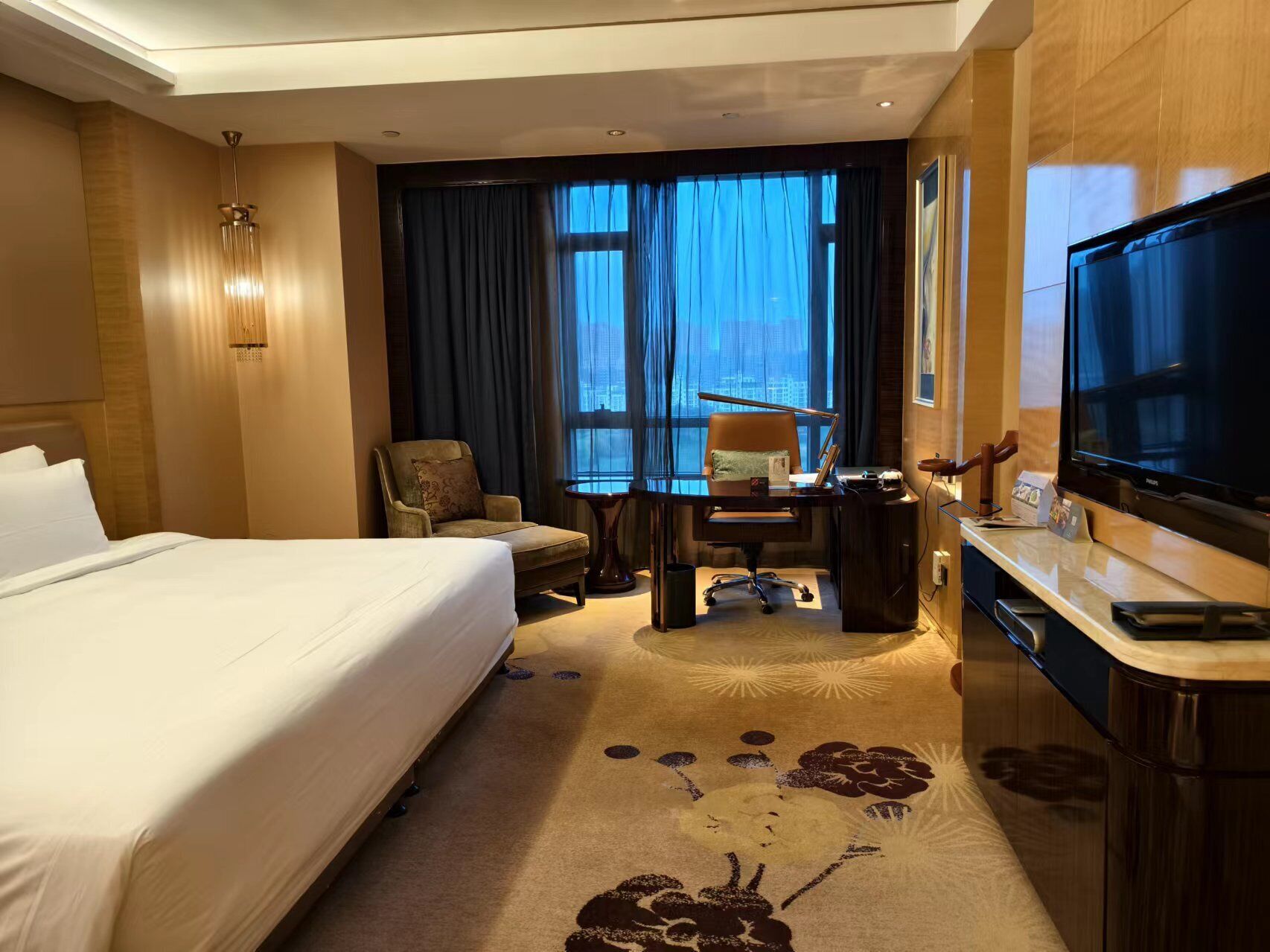 Ein Hotelzimmer mit einem großen Bett, einem Schreibtisch mit Stuhl, einem Fernseher und Blick auf die Stadt durch das Fenster.