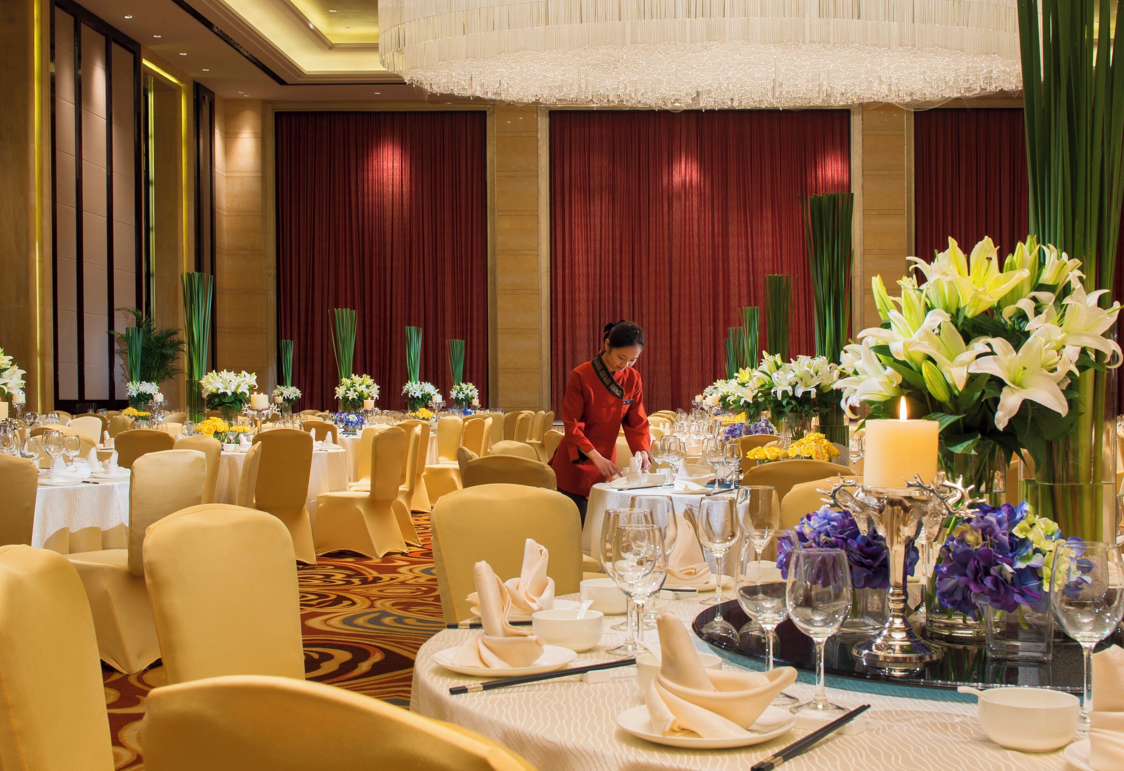 Un salón de banquetes con mesas redondas dispuestas para comer, un miembro del personal atendiendo una mesa y arreglos florales decorativos.