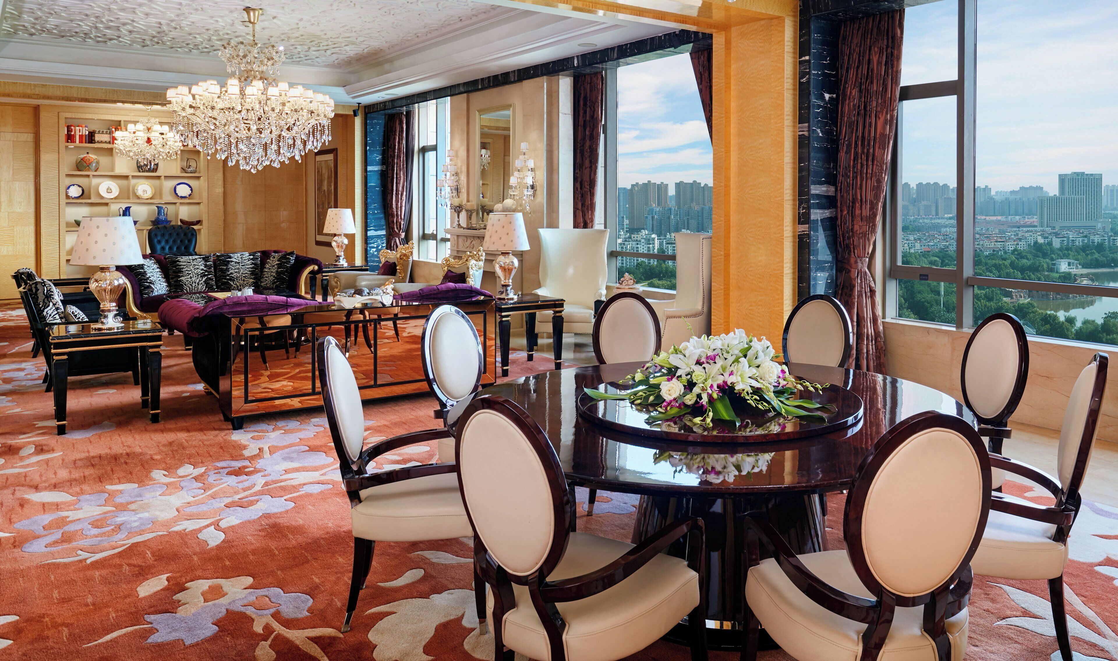 A spacious hotel room with a large dining table, comfortable seating, a chandelier, and a city view.
