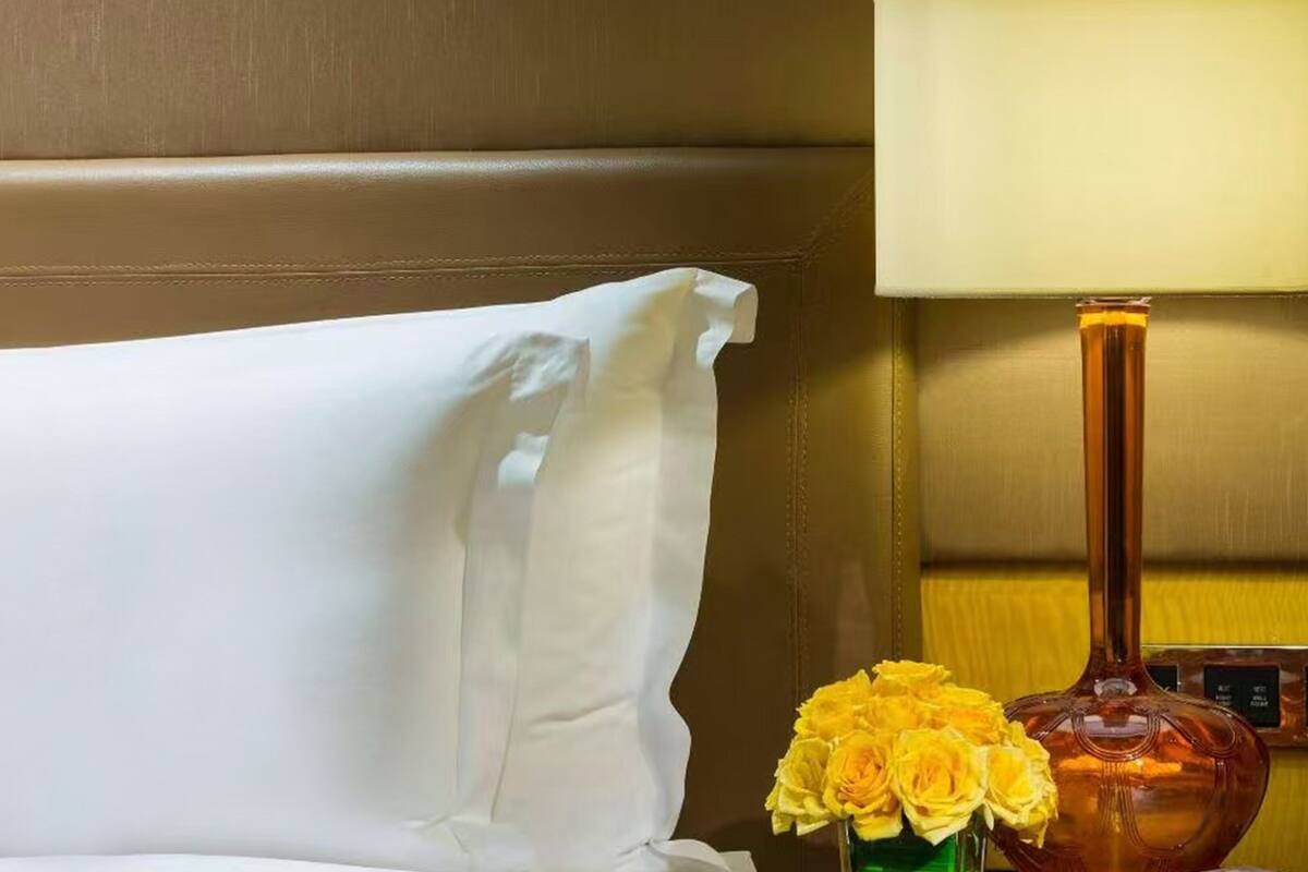 Een hotelkamer met een wit kussen, een vaas met gele bloemen en een lamp.