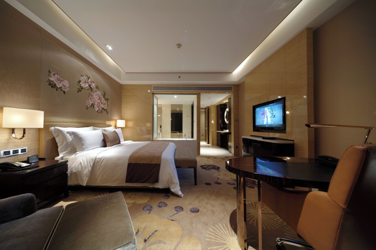 Hotelkamer met een bed, nachtkastje, lamp, televisie, bureau, stoel en een vloerbedekking.