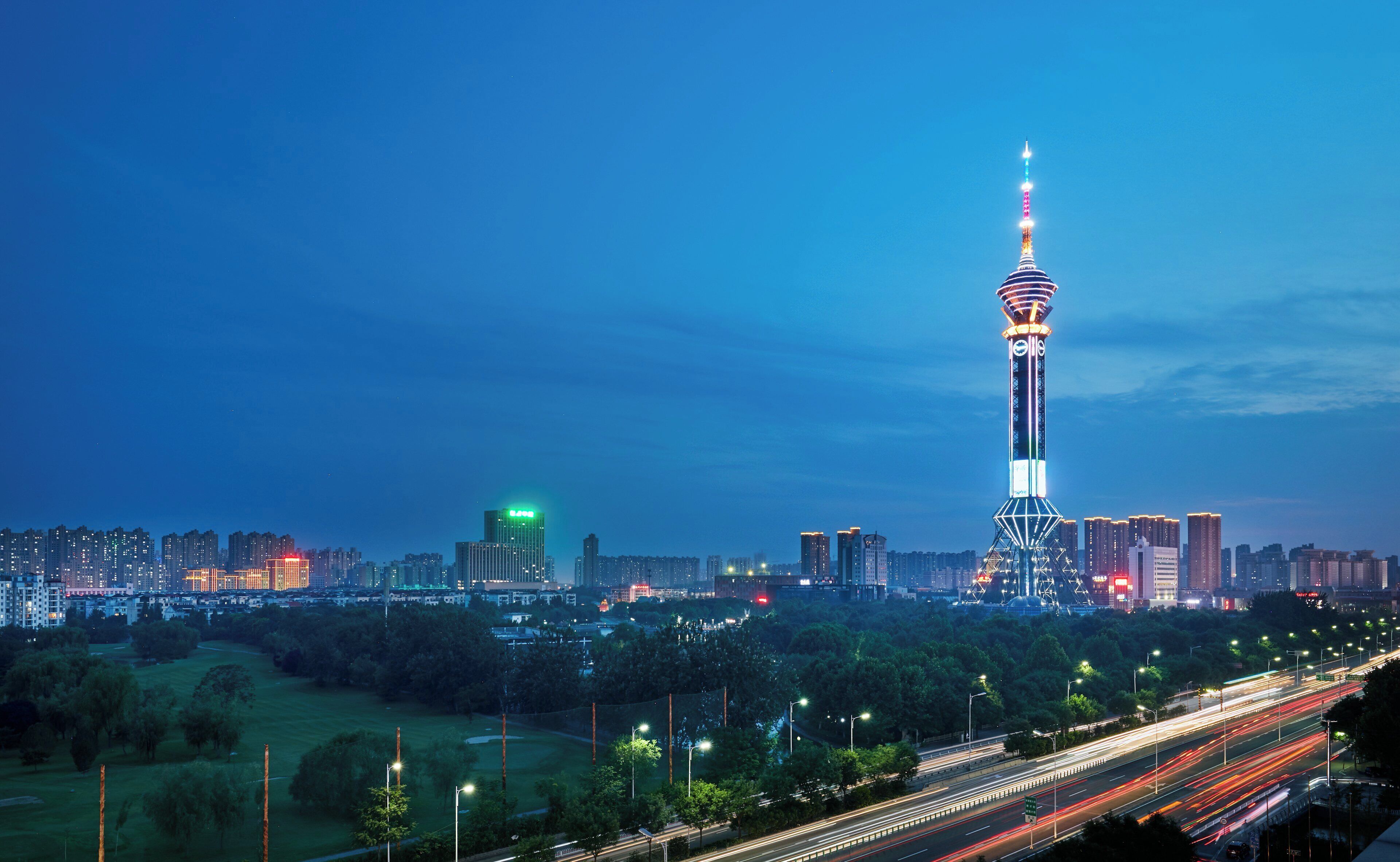 Un paisaje urbano al atardecer con una torre destacada, una autopista con estelas de luz y un parque.