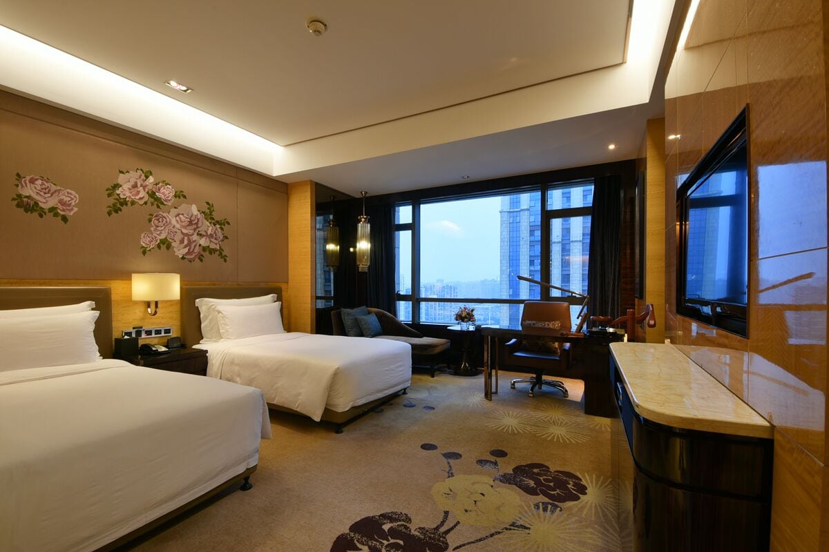 A hotel room with two beds, a desk, a chair, a TV, and a large window offering a city view.