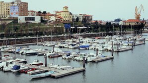 Un puerto deportivo con numerosas embarcaciones amarradas, un puente al fondo y edificios a lo largo de la costa.