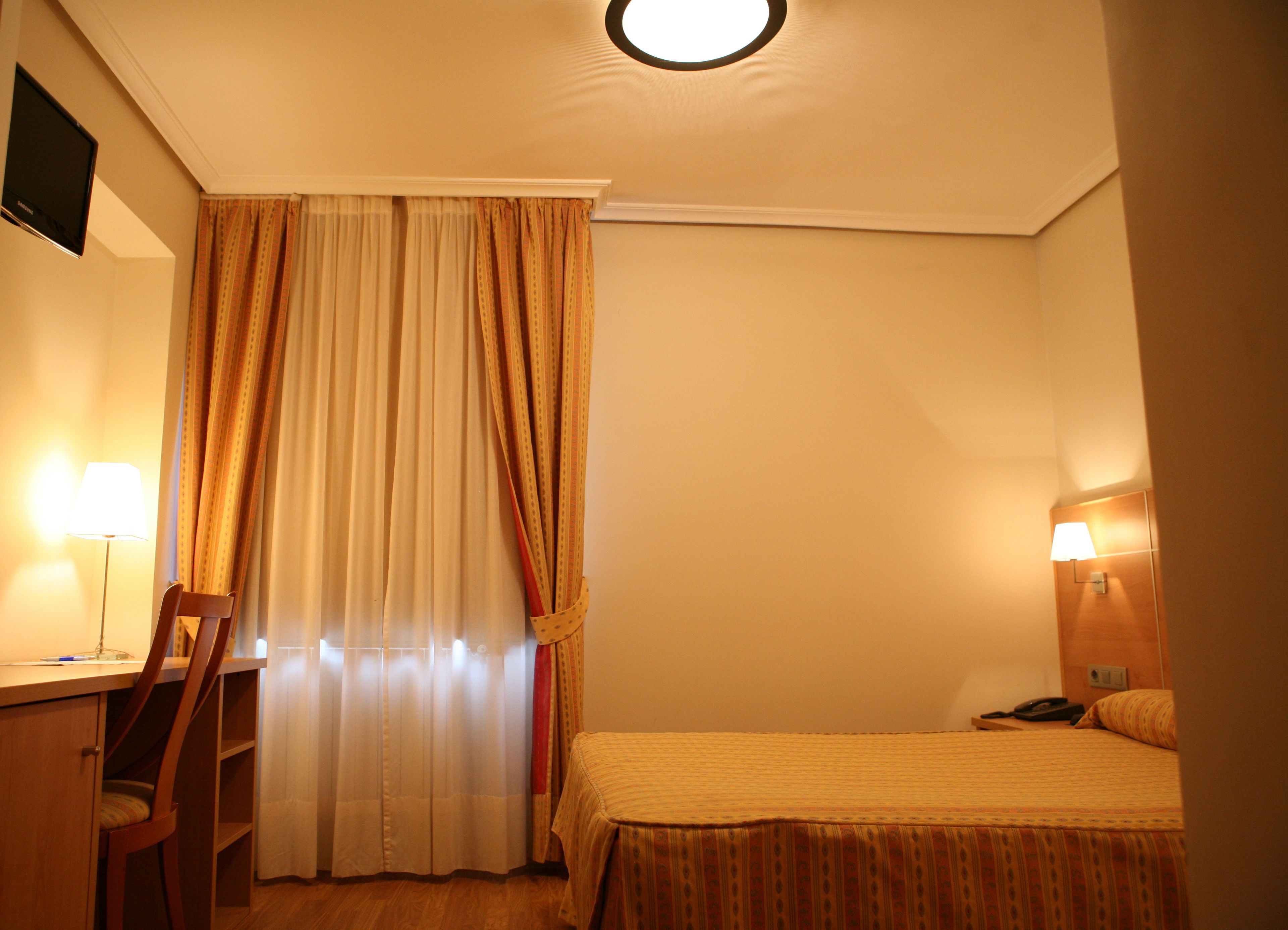 Ett hotellrum med en säng, ett skrivbord, en stol, en lampa och en gardin.