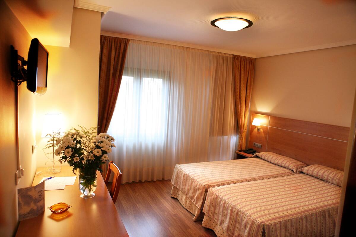 A hotel room with two beds, a desk with a vase of flowers, a lamp, and a television mounted on the wall.