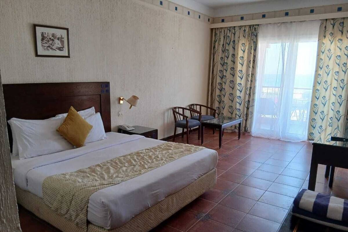 Habitación de hotel con una cama, una mesita, una silla y un balcón con cortinas.