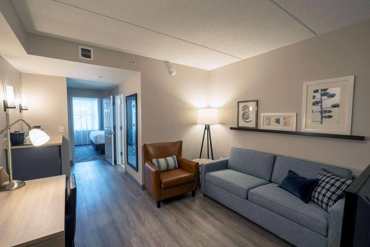 A modern hotel room with a sofa, armchair, desk, lamp, and artwork on the wall.