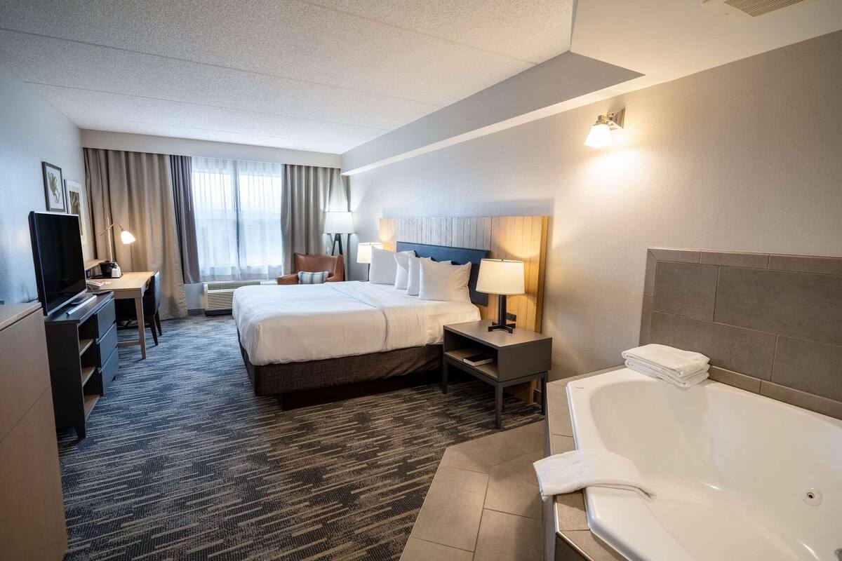 A hotel room with a large bed, a desk with a computer, a TV, a bathroom with a bathtub, and a window with curtains.